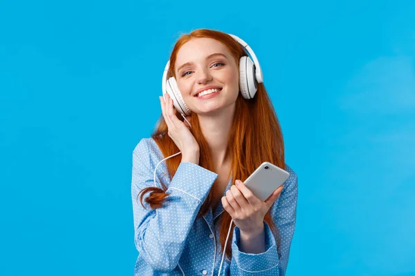 Concepto de tecnología, música y entretenimiento. Alegre atractiva pelirroja caucásica mujer escuchando música en los auriculares, sosteniendo el teléfono inteligente, sonriendo encantado, de pie fondo azul — Foto de Stock
