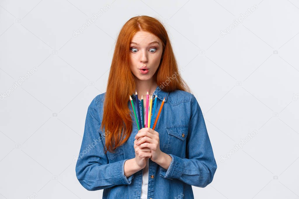 Girl eager start new art course, learn how to design, make models, standing with colored pencils, folding lips amused, looking at tools wondered and thrilled, drawing over white background