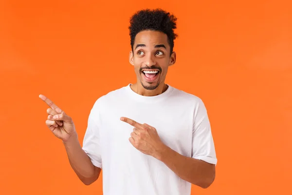 Animado, feliz sorrindo homem afro-americano em t-shirt branca, olhando maravilhado e surpreso com o incrível advrtisement produto, apontando canto superior esquerdo vendo grandes descontos, fundo laranja — Fotografia de Stock