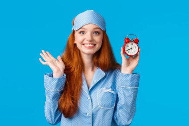 Neşeli ve coşkulu kızıl saçlı kız uyuyakalmak, çalar saati kurmak, kırmızı saati tutmak ve neşe içinde gülümsemek üniversite sınavı için erken kalkmak, gece elbisesi mavi arka planda durmak istemez.