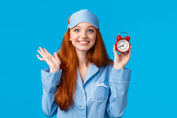 Menina ruiva alegre e entusiasta não quer dormir demais, definir despertador, segurando relógio vermelho e sorrindo alegremente, acordar cedo para o exame universitário, de pé em roupa de dormir fundo azul — Fotografia de Stock