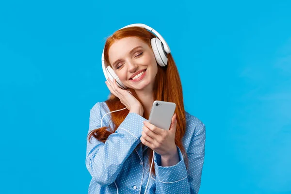 Ternura, mujeres y concepto de estilo de vida. Alegre tierna pelirroja hembra en pijama, cabeza inclinada disfrutando escuchando música, sosteniendo el smartphone y sonriendo como tocando los auriculares de la delicia — Foto de Stock