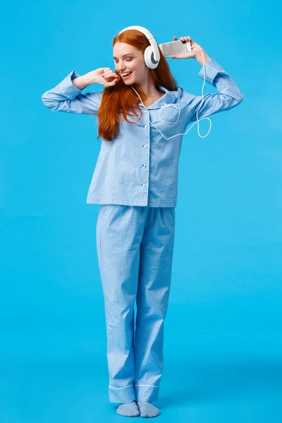 Full-length vertical shot cheerful and upbeat, carefree redhead teenage girl dancing and enjoying beautiful morning, wearing nightwear skipping college stay home listen music in headphones