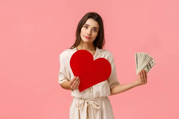 Decisions, relationship and life concept. Alluring romantic young woman having decision made, holding heart card near chest and showing money, tilt head smiling, making choice, pink background