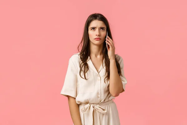 Ernsthafte und gestresste junge brünette Frau, die etwas Seltsames hört, wartet jemand, der das Telefon abholt, steht über rosa Hintergrund im Kleid, hält das Smartphone in Ohrennähe, rosa Hintergrund — Stockfoto
