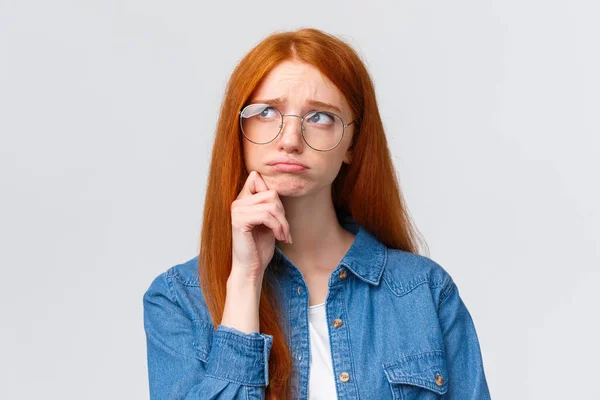 Uneasy sulky cute timid redhead college girl in glasses, look up thoughtful and dreamy, pouting unhappy feeling sad, regret comming party, standing white background indecisive and upset — Stock Photo, Image
