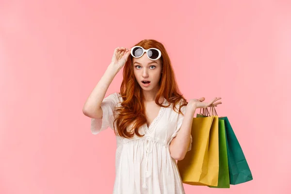 Qué audacia. Sorprendido y sin palabras, asombrado chica pelirroja gafas de despegue para ver algo impresionado, caída de la mandíbula, jadeando, llevar bolsas de compras detrás de la espalda, de pie fondo rosa —  Fotos de Stock