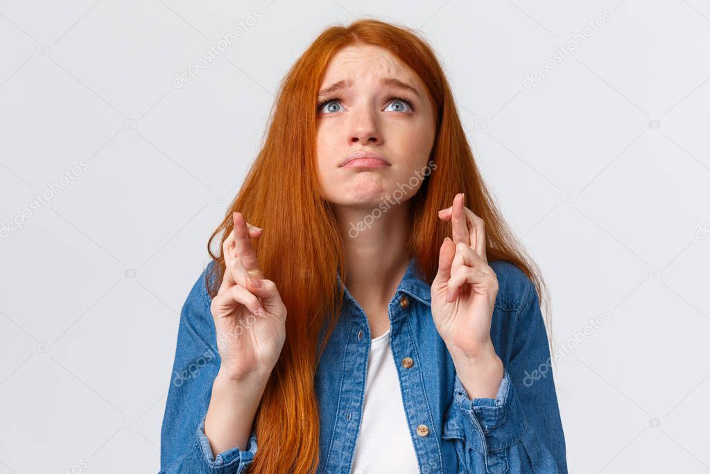Waist-up portrait hopeful and nervous cute gloomy girl with red hair, blue eyes, begging god help, looking at sky with fingers crossed, supplicating, making wish standing white background praying