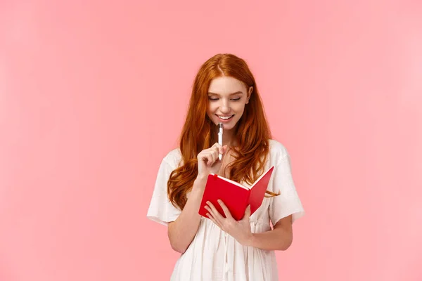 Thoughtful and happy, smiling pretty redhead female in white dress, touching chin with pen while thinking, making grocery or to-do list, writing something in cute red notebook, pink background — Stock Photo, Image