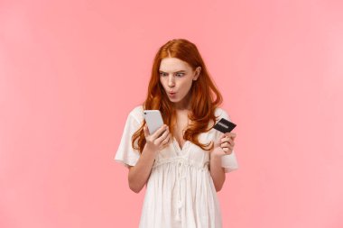 Sinirli ve kızgın, kafası karışmış kızıl saçlı kadın akıllı telefon görüntüsünden memnun değil, bir şeylerin yanlış gittiğinden şikayet ediyor, banka hesabını boş kontrol ediyor, elinde kredi kartı tutuyor.