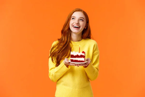 Dreamy happy girl celebrating birthday, having fun, laughing and gazing upper left corner thoughtful, making wish as blowing out candle on delicious, tasty piece b-day cake, orange background — Stock Photo, Image