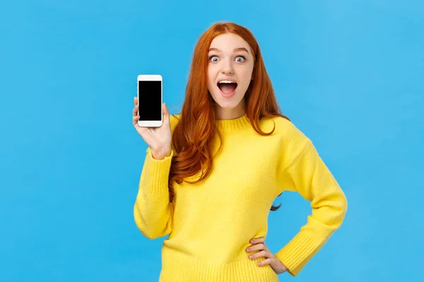 穿着黄色毛衣的红头发女性兴奋地推出了新的应用程序，展示了智能手机的展示，笑着让人着迷地张开嘴逗乐，看相机，蓝色背景 — 图库照片