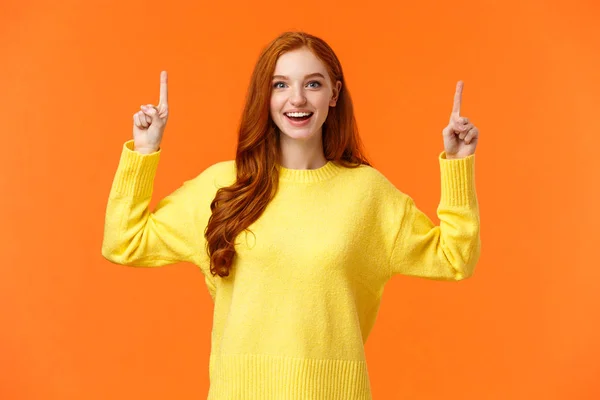 Emocionado, tierna chica pelirroja guapa con pecas, usar suéter amarillo, apuntando hacia arriba y sonriendo alegremente, contando sobre la promoción, publicidad producto superior, fondo naranja —  Fotos de Stock