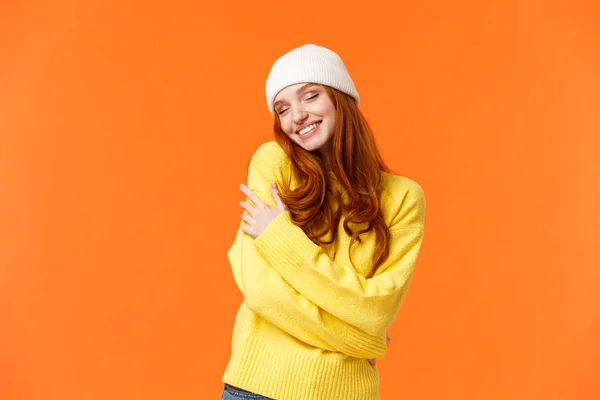 Conceito de ternura, férias de inverno. Mulher ruiva romântico encantador em suéter macio, gorro branco, fechar os olhos e abraçar o próprio corpo, abraçando-se, sorrindo alegre, fundo laranja — Fotografia de Stock