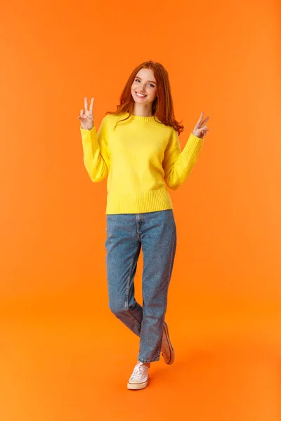 Vertical pleine longueur mignonne belle rousse adolescente en jeans et pull jaune d'hiver debout avec des signes de paix sur fond orange, souriant posant exprimer des émotions de joie insouciantes — Photo