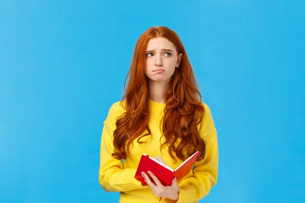 Troubled and pensive uneasy pretty redhead female, pouting and frowning looking away thoughtful, feeling sad and distressed, reading something bad in notebook, standing blue background — Stock Photo, Image