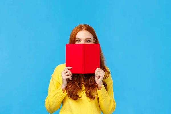 Sassy et créatif heureuse rousse charismatique femme, adolescent cachant visage derrière carnet rouge et souriant, souriant sournoisement, cacher à un ami ce qu'elle a écrit dans son journal, debout fond bleu — Photo