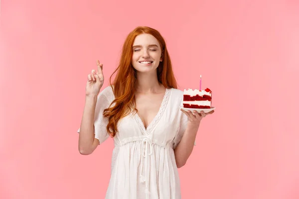 Celebration, happiness concept. Attractive dreamy redhead girl in lovely white dress, holding b-day cake, cross finger good luck, bite lip tempting close eyes and making wish to blow candle — Stock Photo, Image