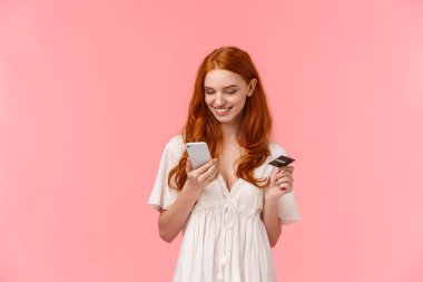 Alışveriş, kolay ödeme ve insanlar konsepti. Beyaz elbiseli güzel bir kız, elinde akıllı telefon ve kredi kartı ile online mağazada hediye seçiyor, teslimat için internet üzerinden ödeme yapıyor, pembe arka plan