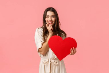 Aşk, güzellik ve mutluluk konsepti. Şehvetli, çekici, seksi elbiseli genç bir kadın, dudaklarına dokunuyor şehvet veya cazibe ifade ediyor, gülümsüyor, bir şeyler denemek istiyor, kırmızı büyük bir kalp tutuyor, sevgililer günü.