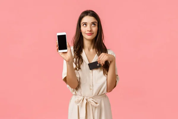 Sonhador e coquete boa aparência jovem mulher no vestido, sonhando esperando ordem de entrega on-line, segurando smartphone e cartão de crédito, mostrando tela móvel, de pé fundo rosa — Fotografia de Stock