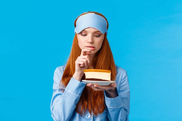 Indecisive redhead girl cant resist temptation, biting finger and looking with desire at slice cake, want eat dessert but worry calories, standing blue background in nightwear and sleep mask