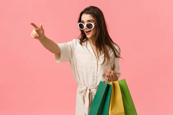 Sassy and elegant young woman in dress, seeing exactly what need, holding shopping bags, pointing finger sideways and looking determined camera, standing pink background excited — Stock Photo, Image