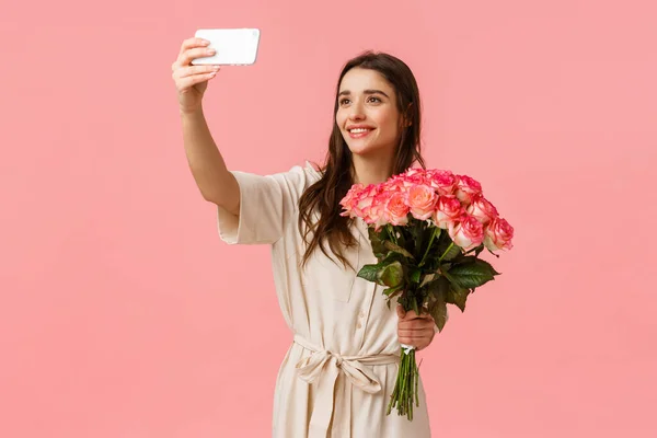Birthday, holidays, romance concept. Charming alluring young caucasian woman receive delivery, got gift flowers, taking selfie on smartphone with beautiful bouquet, standing happy pink background