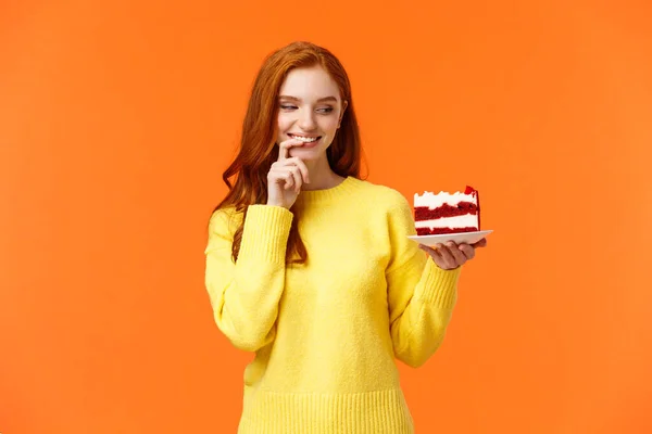 Cheerful and satisfied, happy redhead woman have cheat-day eating delicious food, holding tasty piece cake, biting lip and smiling, cant resist temptation, desire take bite, orange background — Stock Photo, Image