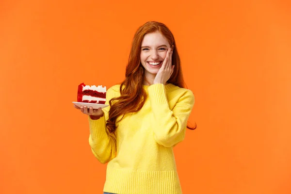 Happy lovely caucasian redhead female holding tasty cake with cream, touch cheek as receive congratulations being birthday girl, celebrating b-day with family, standing orange background — Stock Photo, Image