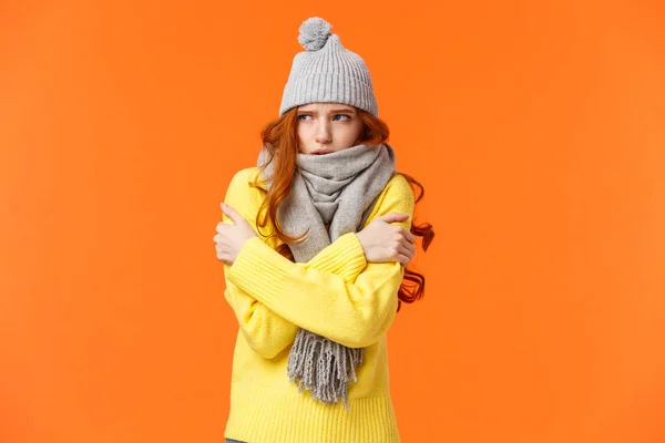Its freezing cold outside. Cute redhead girl shaking and embracing herself to warm up, waiting someone on street in grey scarf, winter hat during snow and low temprature weather, orange background