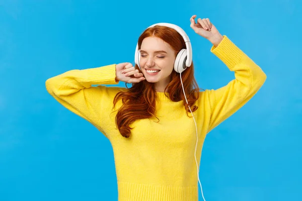 Despreocupado encantadora pelirroja hembra en suéter amarillo, bailando relajado y alegre con las manos levantadas, los ojos cerrados y sonriendo feliz, escuchar auriculares de música, de pie fondo azul — Foto de Stock