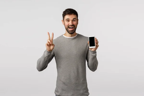 Friendly and carefree funny bearded gay man in grey sweater edit profile picture online dating app, make peace sign, showing smartphone display and smiling happily, standing white background — Stock fotografie