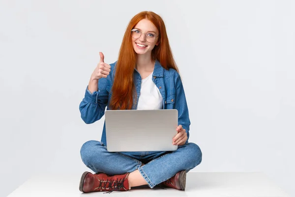 Education, internet and people concept. Female redhead digital nomad, freelance girl in glasses, finished project before deadline, sitting on floor with crossed legs and laptop, show thumb up