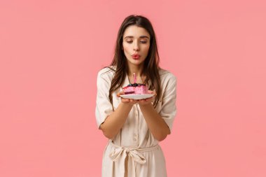 Şehvetli, muhteşem esmer beyaz kız doğum gününü kutluyor, elinde bir tabak pasta ve sönmüş mum tutuyor, dilek diliyor ve neşeyle gülümsüyor, pembe arka planda kaygısız duruyor.