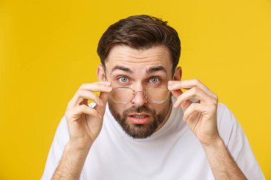 Genç yakışıklı bir adamın kameraya bakıp komik yüzünü göstermesi, gözlük takması. Sarı arkaplanda izole görünüm.