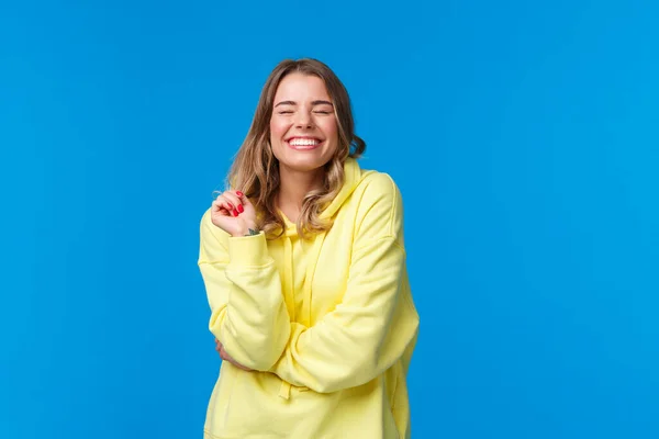 Χαρούμενα ανέμελη ευχαριστημένος κορίτσι αισθάνονται ευτυχείς και ενθουσιασμένοι, κοντά μάτια ονειρικό χαμόγελο με τέλειο ακτινοβολούν χαμόγελο, αφή σκέλος των μαλλιών, στέκεται σε κίτρινο hoodie πάνω από το μπλε φόντο — Φωτογραφία Αρχείου