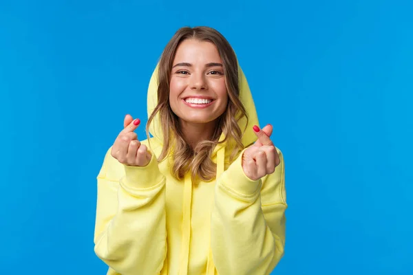 Primer plano retrato de linda y tonta chica rubia caucásica en sudadera con capucha amarilla, mostrando el gesto del corazón coreano y sonriendo, vibing, disfrutando de la fiesta, divertirse sobre fondo azul —  Fotos de Stock