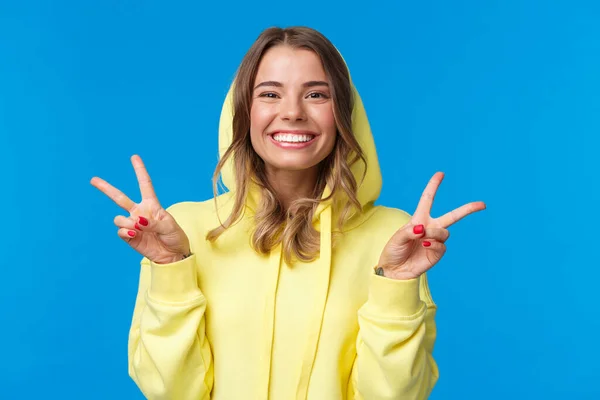 Primer plano retrato lindo tonto rubio europeo hembra en sudadera con capucha amarilla, mostrando gesto de paz y optimista sonriente, enviar vibración positiva, de pie fondo azul optimista, hacer pose kawaii —  Fotos de Stock