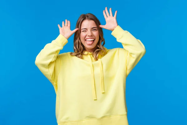 Porträtt av lockande bekymmerslös kaukasiska blond flicka har roligt, visar fånga mig om du kan, lekfull grimace med händerna nära huvudet, stick tunga och leende, stående blå bakgrund — Stockfoto