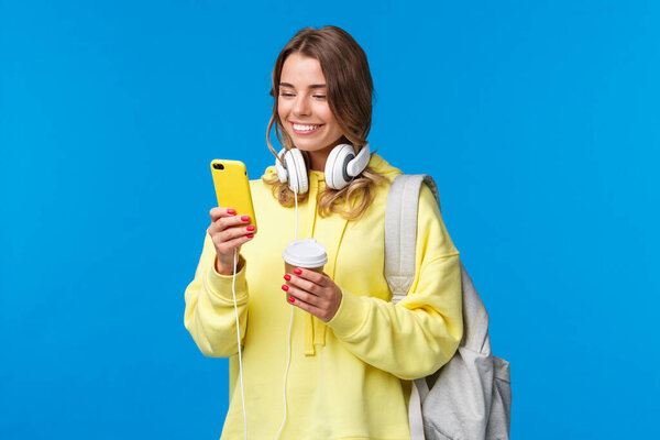 Girl using mobile phone to contact friend after college, texting as walking along street with backpack, take-away coffee cup and headphones, smiling joyfully smartphone display