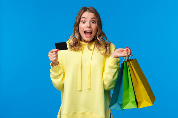 Взволнованная блондинка готова потратить все деньги на свой банковский счет, кричать взволнованно и радостно во время покупок, держа сумки с товарами и кредитной картой, стоять на синем фоне
