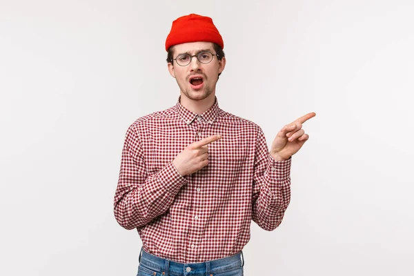 Waist-up portrait unimpressed reluctant and bored young man in glasses and red beanie yawning uninterested as pointing upper right corner at something that not worth attention, white background — ストック写真
