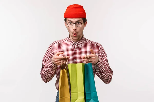 Waist-up portrait excited and surprised handsome young man with moustache, wear glasses and beanie, looking amazed camera say wow as open shopping bags with gifts, stand white background