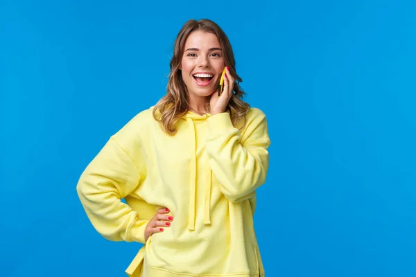 Despreocupada chica rubia europea guapa en sudadera con capucha amarilla animada hablando en el teléfono móvil, cámara sonriente como mesa de reserva para amigos en el bar, orden de entrega de alimentos, fondo azul —  Fotos de Stock