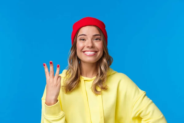 Mulher caucasiana de boa aparência em gorro vermelho e capuz amarelo, mostrando o número três e sorrindo, faça a ordem ou reserva, de pé orgulhoso e encantado, fundo azul — Fotografia de Stock