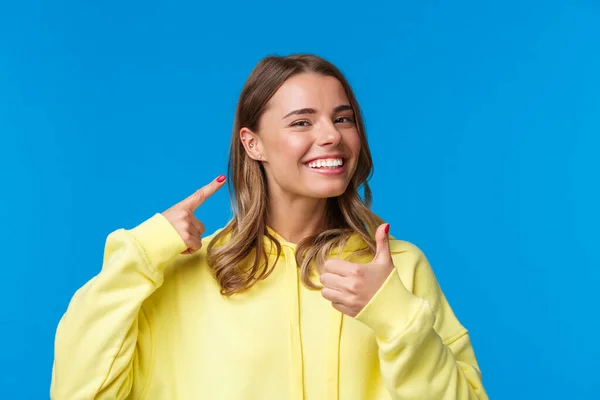 Alegre y complacida, satisfecha y atractiva hembra europea apuntando a la oreja perforada y sonriendo encantada, muestra los pulgares hacia arriba como sus nuevas joyas, de pie fondo azul —  Fotos de Stock