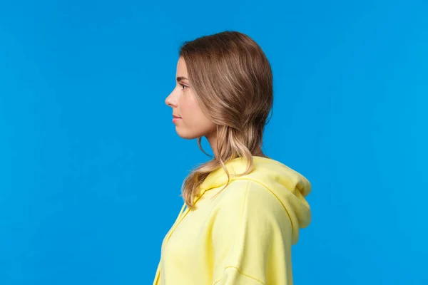 Close-up profile portrait of serious-looking attractive young caucasian female with short blond hair, looking left, standing in yellow hoodie, standing blue background straight — Stock Photo, Image