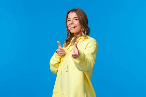 Oye, tú. Cheeky y despreocupado linda chica europea moderna en sudadera con capucha amarilla, guiño y hacer el gesto de pistolas de dedo en la cámara para decir hola, concepto de saludo informal. fondo azul —  Fotos de Stock