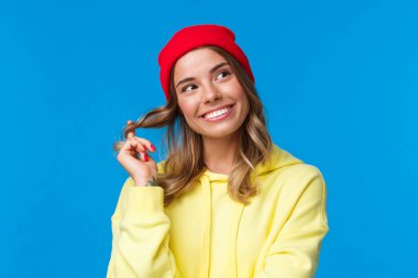 Close-up portrait dreamy silly cute european female in red hipster beanie, yellow hoodie, rolling curl on hair and gazing upper right corner thoughtful, standing blue background